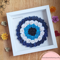 Cuadro de ojo turco hecho con flores de papel blancas, azules y negras, con marco blanco que resalta el color de las flores. Este diseño aportará armonía y elegancia a tus espacios.