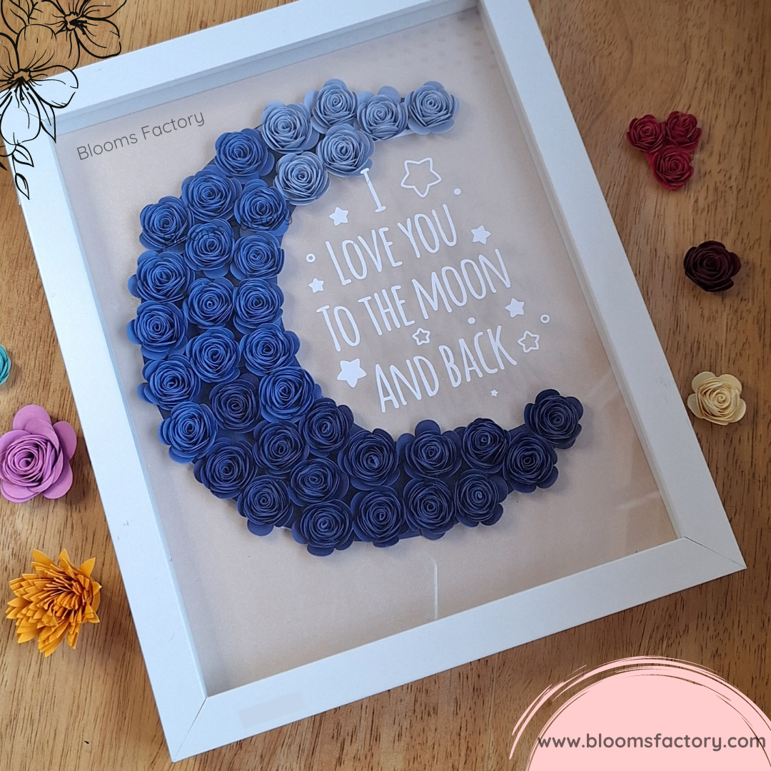 Cuadro decorativo con una luna creciente hecha de flores de papel azules, enmarcada en blanco. El fondo tiene el texto "I LOVE YOU TO THE MOON AND BACK" con pequeñas estrellas.