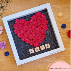 Bloom Box con diseño en forma de corazón de rosas de papel de color rojo vibrantes enmarcado en un cuadro blanco, con la palabra ‘MAMA’ escrita con fichas de madera debajo, sobre un fondo oscuro con brillo. 