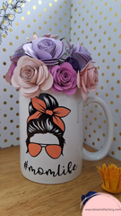 Taza blanca con diseño de mujer con moño y gafas de sol y hashtag #momlife, llena de flores de papel de colores y el #momlife