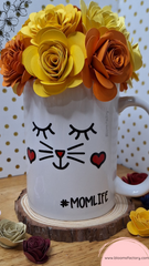 Taza decorativa coronada con flores de papel, la base está decorada con un rostro adorable de ojos tiernos y mejillas sonrosadas acompañada de unos bigotes de gato que te hará sonreír cada vez que la veas. 