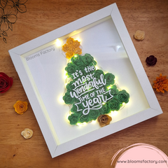 Un cuadro decorativo que presenta un árbol de Navidad hecho con flores de papel verde, con una estrella dorada y el texto ‘It’s the most wonderful time of the year’ sobre él y luces que le dan un toque único y original. Es ideal para darle un toque festivo y original a tu hogar, o para sorprender a tus seres queridos con un regalo único y personalizado. 