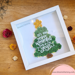 Un cuadro decorativo que presenta un árbol de Navidad hecho con flores de papel verde, con una estrella dorada y el texto ‘It’s the most wonderful time of the year’ sobre él y luces que le dan un toque único y original. Es ideal para darle un toque festivo y original a tu hogar, o para sorprender a tus seres queridos con un regalo único y personalizado. 